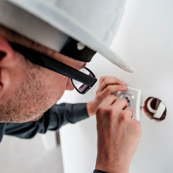 man wearing glasses and a white hard hat works on electrical outlet