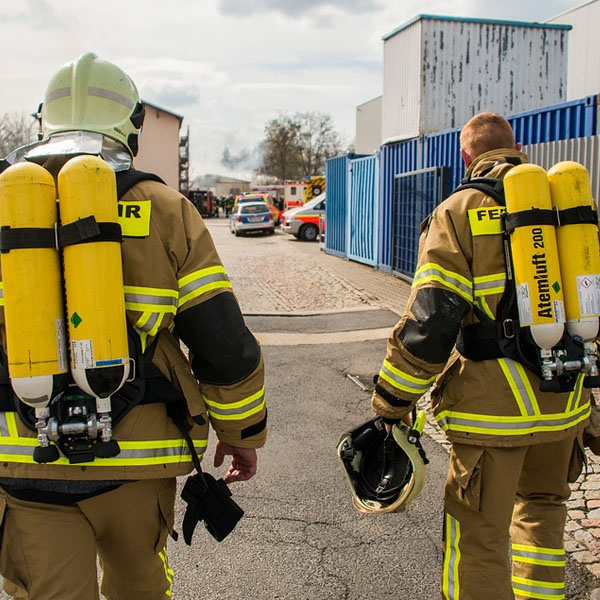the backs of two fireman wearing fire resistant gear and oxygen tanks walk side by side