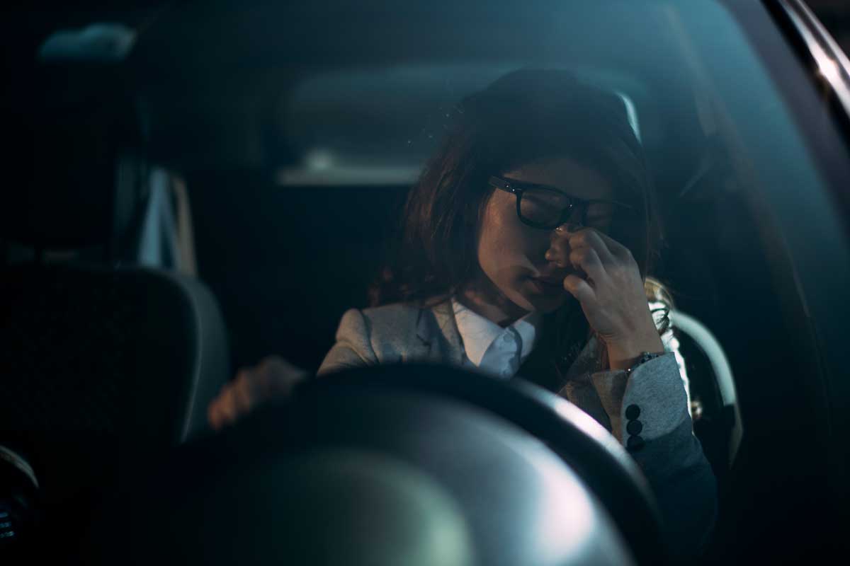 woman with closed eyes sits in driver's seat of car rubbing her eyes with one hand