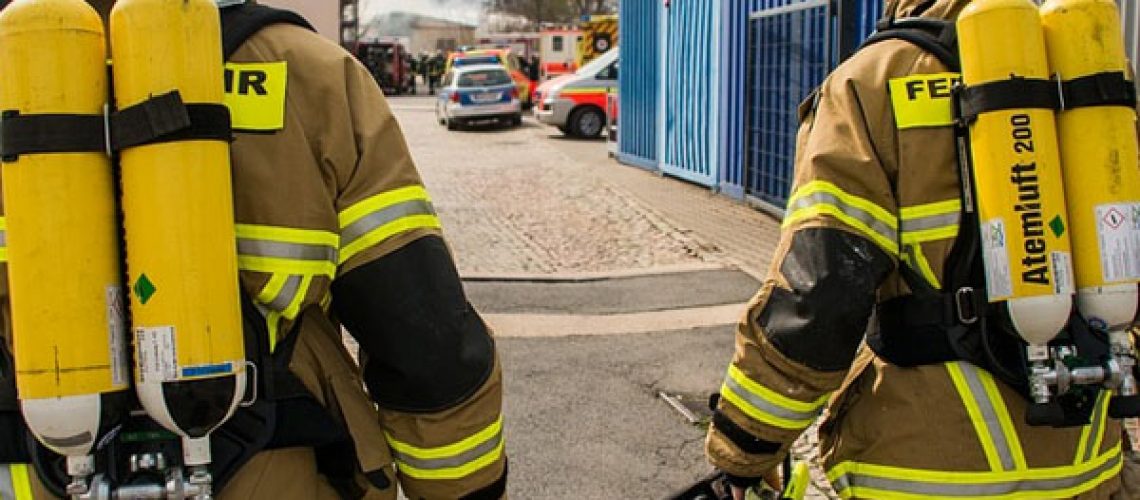 the backs of two fireman wearing fire resistant gear and oxygen tanks walk side by side